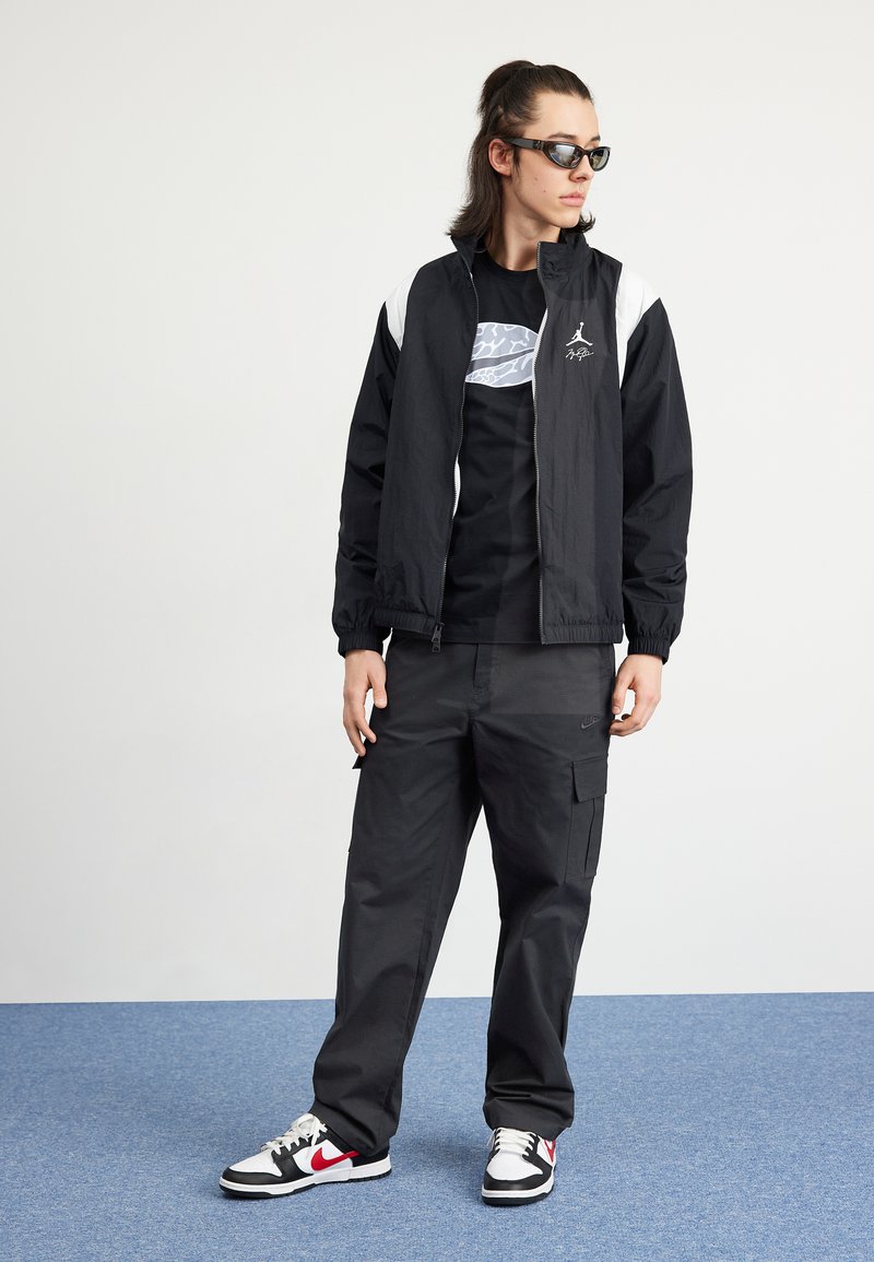Veste légère noire avec des accents blancs, t-shirt noir avec un motif graphique, pantalon cargo noir, et baskets noir et blanc avec un détail rouge.