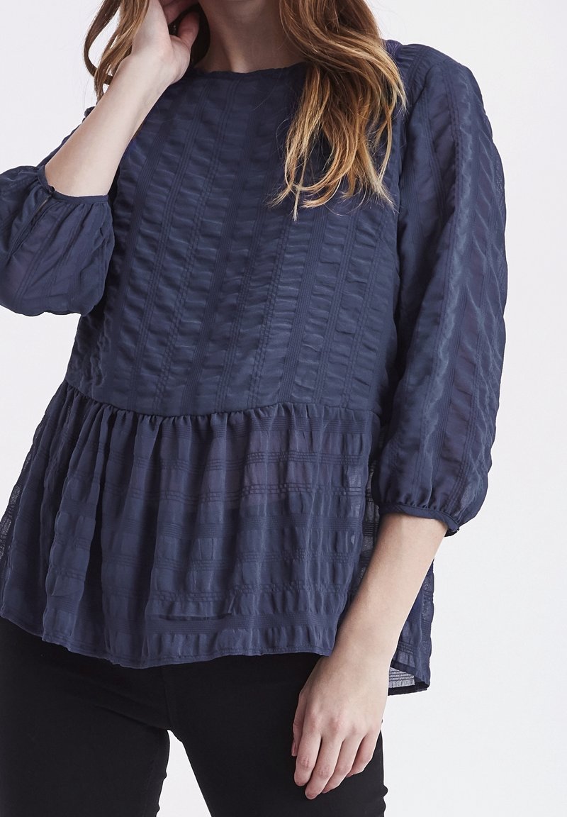Námořnická modrá blůza s texturovanou tkaninou, která má nařasený peplum lem a rukávy tříčtvrtinové délky s elastickými manžetami.