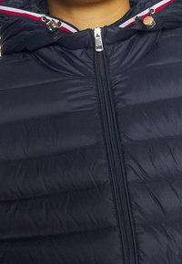 Nærbillede af en navy jakke med lynlås, hætte med rød, hvid og blå kant samt justerbare snore.