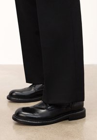 Fekete bőr dresszcipők kerek orral, karcsú kialakítással és varrott részletekkel, fekete bővebb nadrággal párosítva.