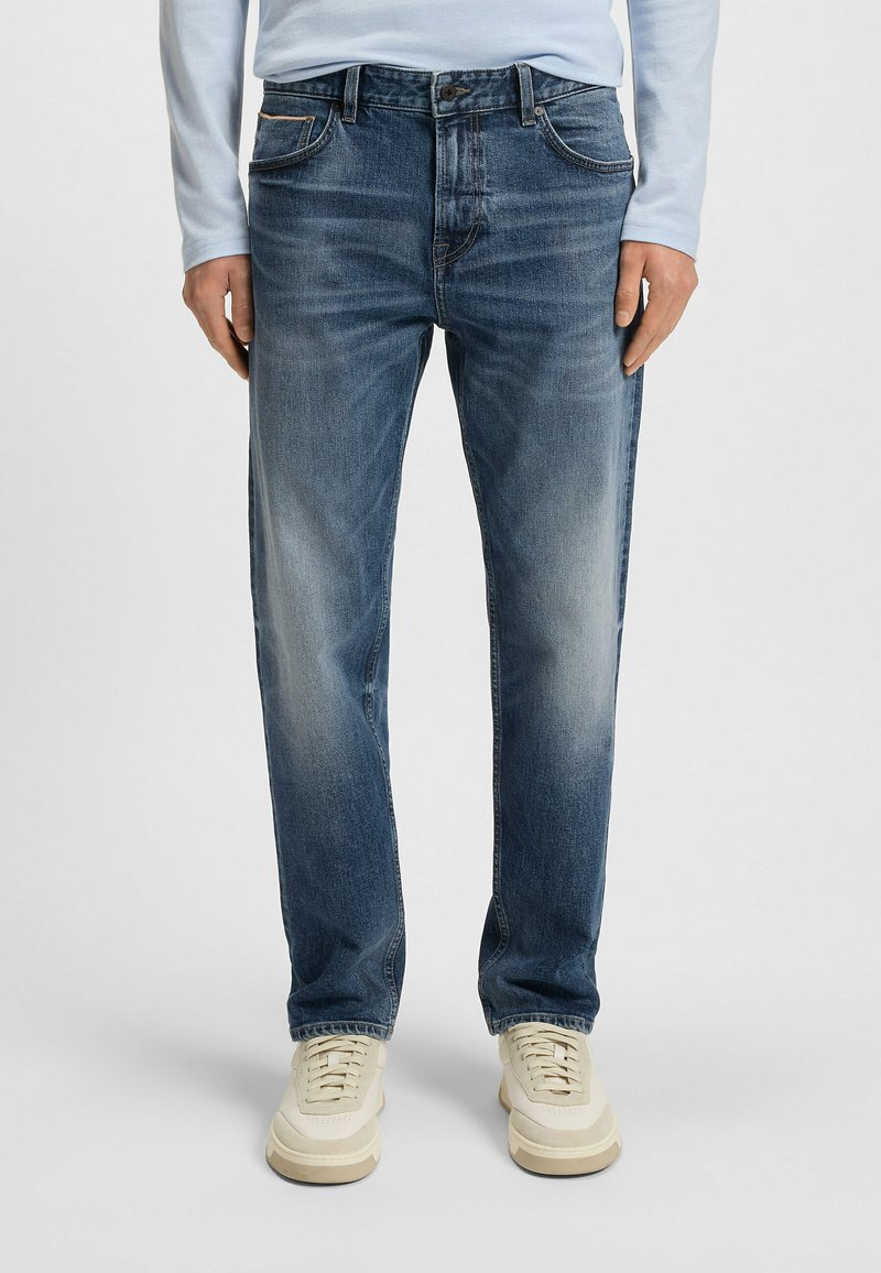 Jeans in denim di un blu scuro con sbiaditure chiare, vestibilità dritta, design a cinque tasche e chiusura con bottone, abbinati a sneakers beige.