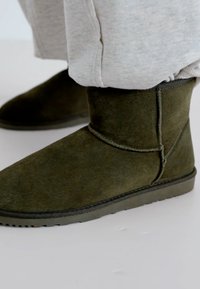 Botines de ante verde oliva con una textura suave, puntera redondeada y suela de goma, que presentan un detalle de costura a lo largo del costado.