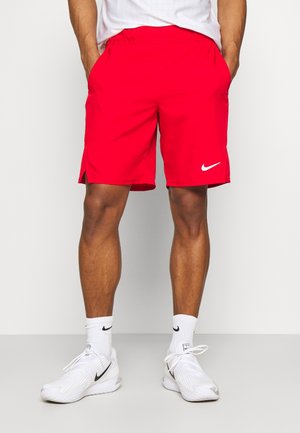 Homme portant un short Nike rouge vif avec des fentes latérales, des chaussettes blanches Nike et des baskets blanches Nike, debout les mains dans les poches.