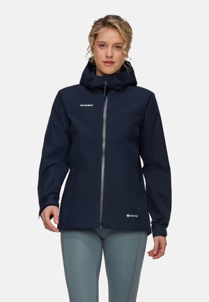 Kobieta ubrana w granatową kurtkę Mammut Gore-Tex z kapturem i zamkiem z przodu oraz jasnoszare legginsy, stoi na jednolitym tle.
