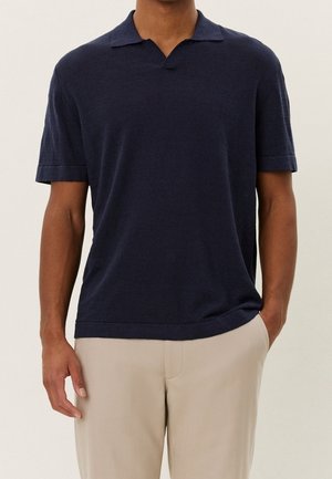 Homme portant un polo bleu marine à manches courtes et un pantalon beige, debout avec une main dans la poche devant un arrière-plan uni.