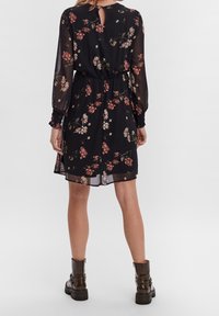 Vestido negro con largas mangas transparentes y un patrón floral en burdeos y beige. Presenta un escote en forma de llave en la parte posterior y una cintura elástica, combinado con botas hasta el tobillo.