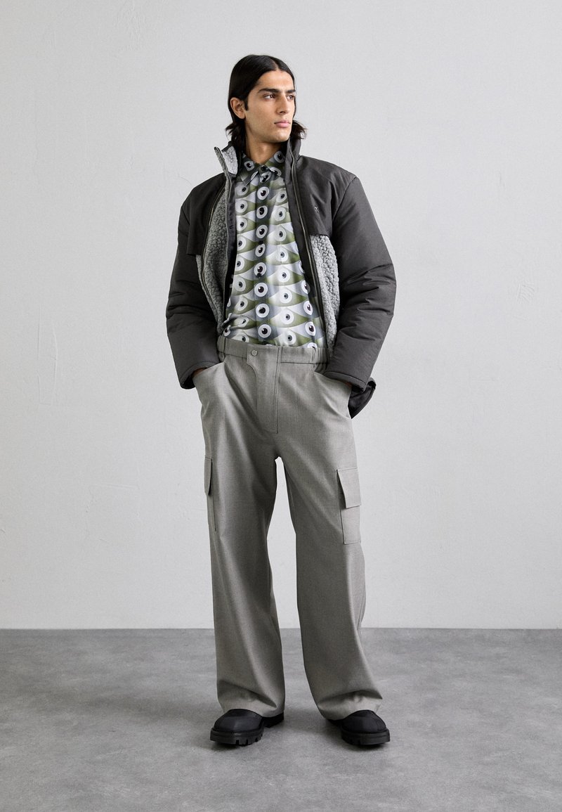 Polstret jakke i mørkegrå med grå fleecefore, mønstret skjorte i grønne og sorte nuancer, samt lysegrå cargo-bukser med lommer. Sorte sko.