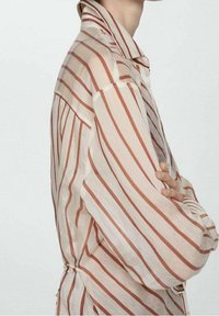 Camicia a righe in tessuto morbido e trasparente. Rigature orizzontali beige e ruggine con colletto, vestibilità rilassata e polsini con bottoni. Lacci laterali regolabili.