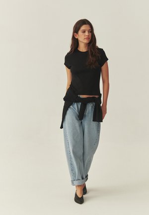 Woman with long brown hair wearing a black fitted t-shirt, light blue rolled-up jeans, black shoes, and a black sweater tied around her waist.