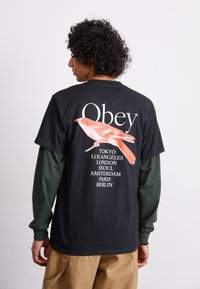 Juodas trumpų rankovių marškinėlis su raudono paukščio grafika ir žodžiu "Obey" viršuje, su miesto pavadinimais išvardytais vertikaliai ant nugaros.