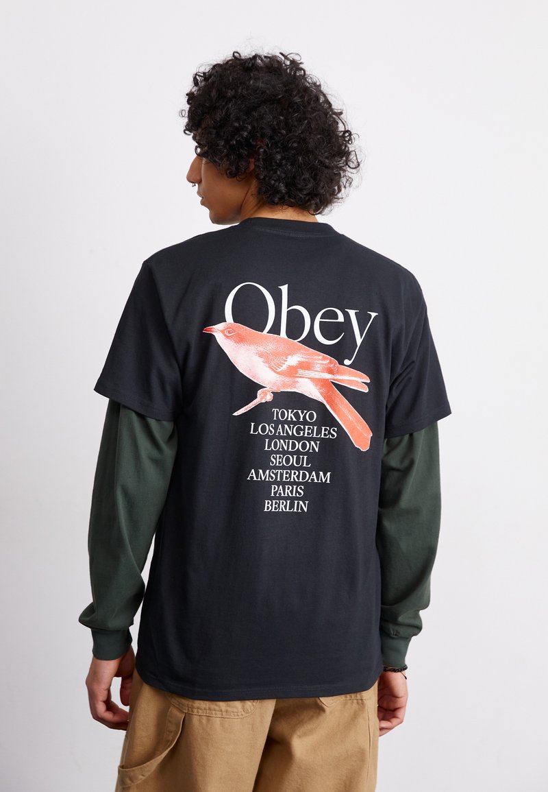 Juodas trumpų rankovių marškinėlis su raudono paukščio grafika ir žodžiu "Obey" viršuje, su miesto pavadinimais išvardytais vertikaliai ant nugaros.