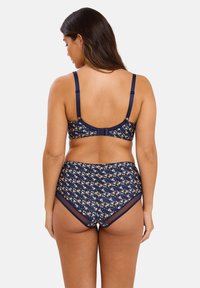 Vrouw met lang haar in een marineblauwe bloemenbeha en bijpassende hoge slip, gezien vanuit de achterkant tegen een effen achtergrond.