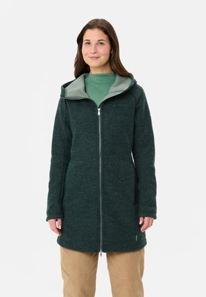 Femme portant une longue veste polaire zippée vert foncé avec capuche et doublure intérieure vert clair, associée à un pantalon beige.