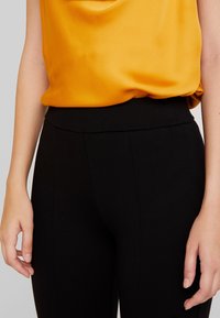 Černé legíny s vysokým pasem spárované s volnou, jasně žlutou saténovou blůzou. Látka má hladkou strukturu a jemný lesk.