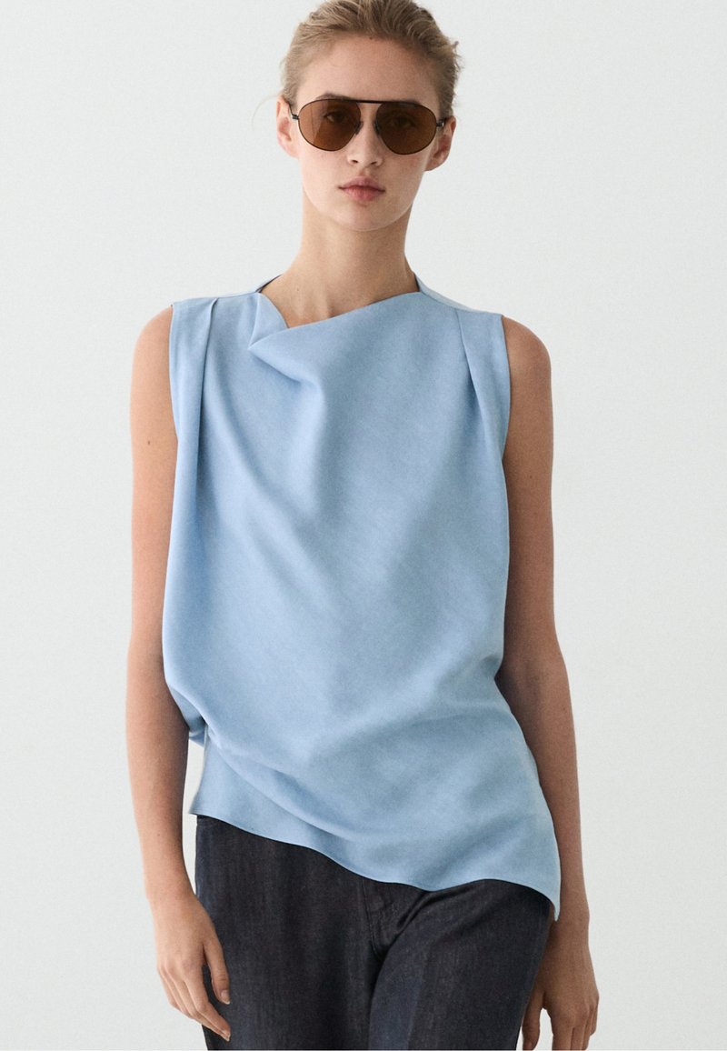 Donna con occhiali da sole marroni, top azzurro chiaro senza maniche con dettaglio piegato e pantaloni scuri, in piedi davanti a uno sfondo chiaro e uniforme.