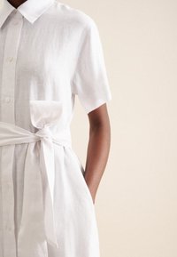 Weißes Leinenhemdkleid mit kurzen Ärmeln, einem Kragen, Knöpfen vorne, einer Tasche und einem gebundenen Taillengürtel, aus strukturiertem Stoff.