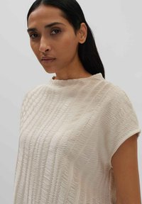 Top beige chiaro con un motivo testurizzato e ondulato. Presenta maniche corte e una vestibilità rilassata, con un colletto alto e una leggera lucentezza.