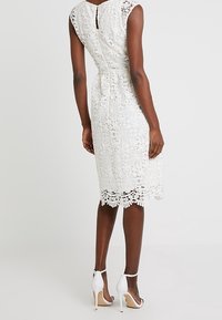 Robe en dentelle blanche avec motif floral, longueur genou, manches courtes, détail dos goutte d'eau, associée à des sandales blanches à talons hauts.