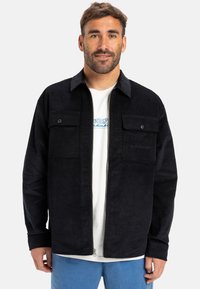 Giacca in corduroy nera con zipper frontale, due tasche sul petto e finitura testurizzata, indossata sopra una maglietta bianca e pantaloni blu.