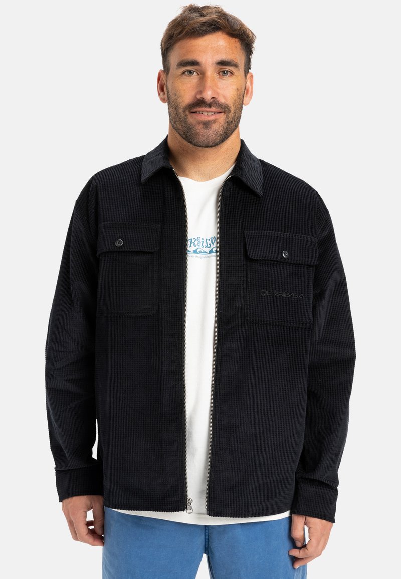 Giacca in corduroy nera con zipper frontale, due tasche sul petto e finitura testurizzata, indossata sopra una maglietta bianca e pantaloni blu.