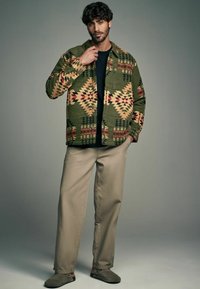 Veste multicolore à motifs géométriques en vert, rouge et beige, portée sur une chemise noire, associée à un pantalon beige et des chaussures gris slip-on.