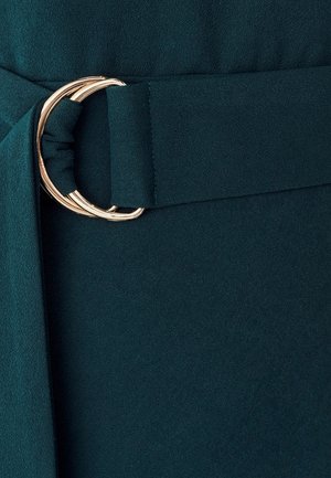 Modrozelená látka s preloženou časťou so zlatým kovovým okrúhlym prackou, vytvárajúca hladkú textúru a štruktúrovaný dizajn.