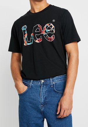 Homme portant un t-shirt noir Lee avec un logo coloré et un jean bleu, debout avec une main dans la poche.