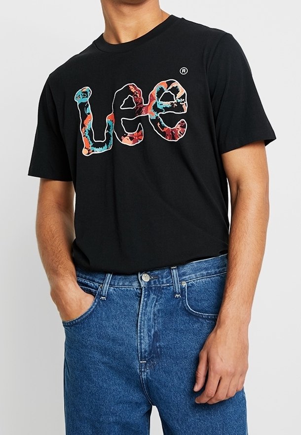Homme portant un t-shirt noir Lee avec un logo coloré et un jean bleu, debout avec une main dans la poche.
