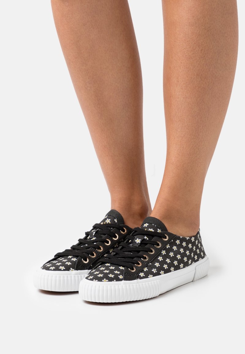 Legs wearing black canvas sneakers with white daisy patterns and thick white rubber soles on a white background.