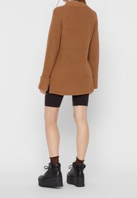 Frau mit einem braunen langen Pullover, schwarzen Radshorts, durchsichtigen braunen Socken und schwarzen Plateauschuhen mit Schnürung, die mit dem Rücken zur Kamera steht.