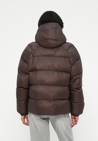 Brun pufferjakke med hætte, med et quiltet design og elastiske manchetter. Stofbutikken har en glat overflade og en let skinnende tekstur.