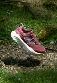 Baskets de sport bordeaux avec une tige texturée, un logo bien en vue et une semelle rembourrée, se détachant sur un fond naturel avec de la verdure.