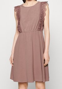 Hihaton pölyruusuvärinen mekko, jossa on joustava vyötärö ja kukkakuvioinen pitsisomistus hartioilla ja sivuilla seisovalla henkilöllä.