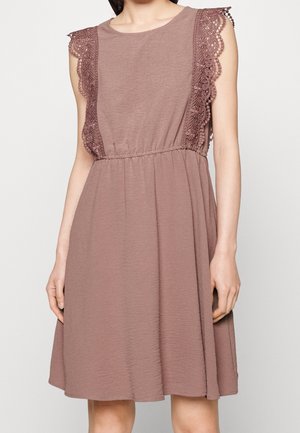 Ärmelloses, staubrosa Kleid mit elastischem Bund und floraler Spitzenverzierung an den Schultern und Seiten bei einer stehenden Person.