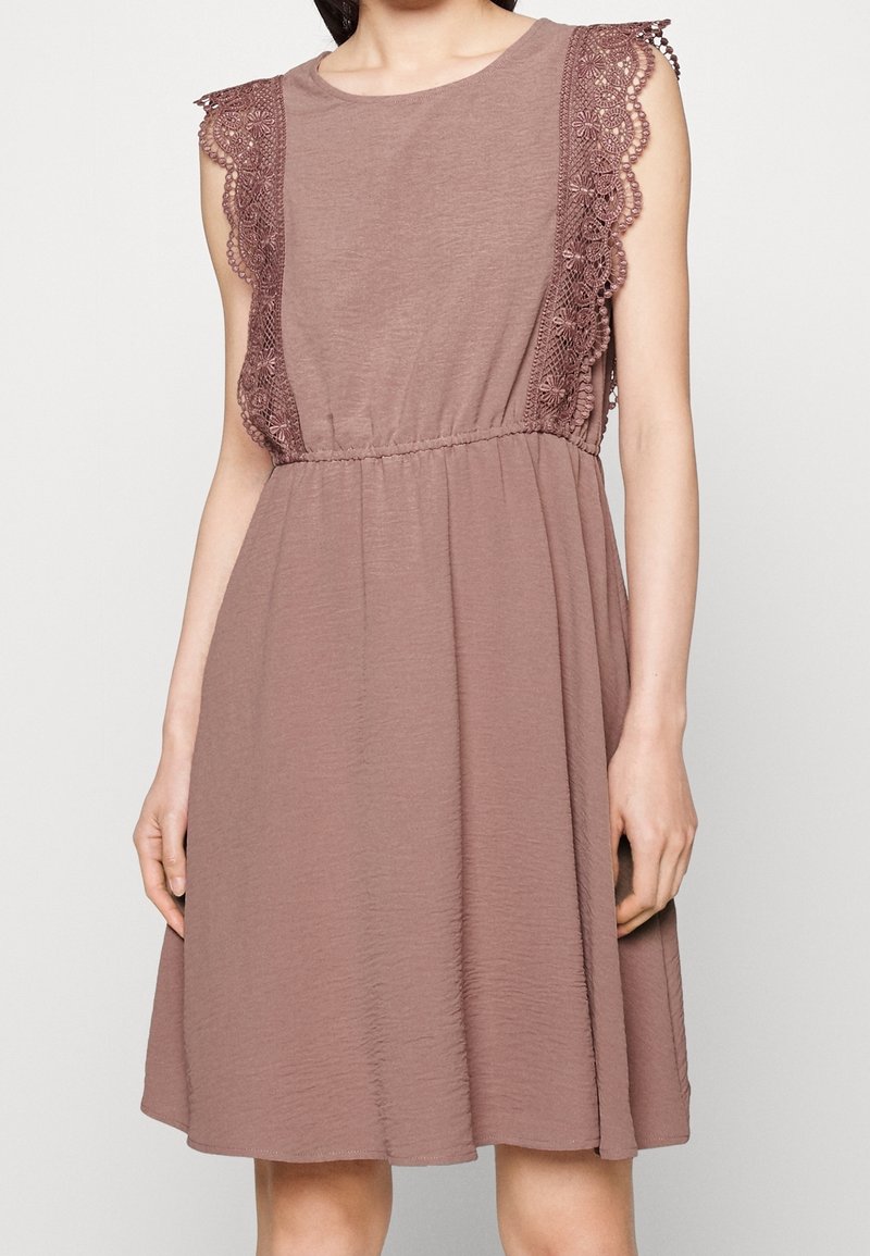 Hihaton pölyruusuvärinen mekko, jossa on joustava vyötärö ja kukkakuvioinen pitsisomistus hartioilla ja sivuilla seisovalla henkilöllä.