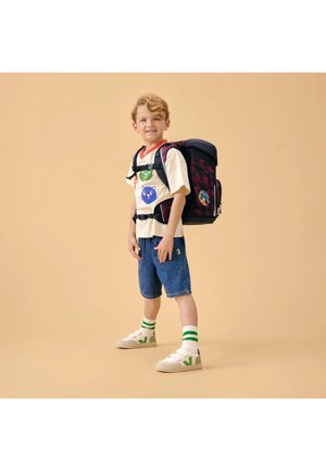 Junger Junge trägt ein T-Shirt mit Katzenmotiv, Jeansshorts und Turnschuhe mit grün gestreiften Socken, der einen großen Rucksack mit Brust- und Hüftgurten trägt.