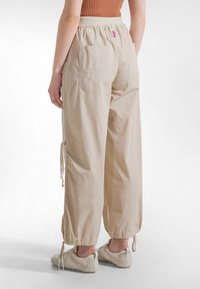 Beige bukser med vide ben, elastisk talje, to baglommer og justerbare snore i anklerne. Fremstillet af letvægtsstof.