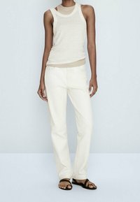 Beige mouwloze top gelaagd over een lichte top, gecombineerd met crème rechte broek en zwarte sandalen. Glad oppervlak overal.