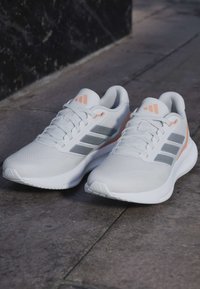 Chaussures de course athlétiques blanches et grises avec des accents pêche et des lacets posées sur un pavé en pierre à l'extérieur.
