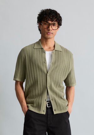 Mann mit lockigem Haar und Brille, der ein olivgrünes, kurzärmliges, geknöpftes Strickhemd über einem weißen Tanktop mit schwarzer Hose trägt.