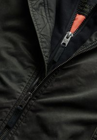 Zwarte bomberjack met een matte afwerking, voorzien van een contrasterende oranje voering, twee ritsen en een gestructureerde stof. Gevoerde details zichtbaar.