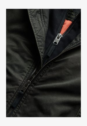 Sort bomberjakke med mat finish, der har en kontrasterende orange foring, to lynlåse og et tekstureret stof. Vatterede detaljer er synlige.