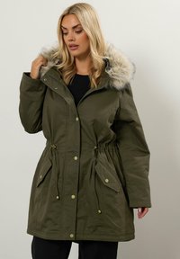 Olivgrön parka med huva i fuskpäls, dragsnör i midjan och fickor med tryckknappar. Har en dragkedjestängning och ett mjukt, texturerat tyg.