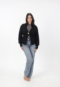 Schwarze Strickjacke mit goldener Knopfleiste, langen Ärmeln und lockerer Passform, kombiniert mit hellblauen, hochgeschnittenen Jeans und durchsichtigen Sandalen.
