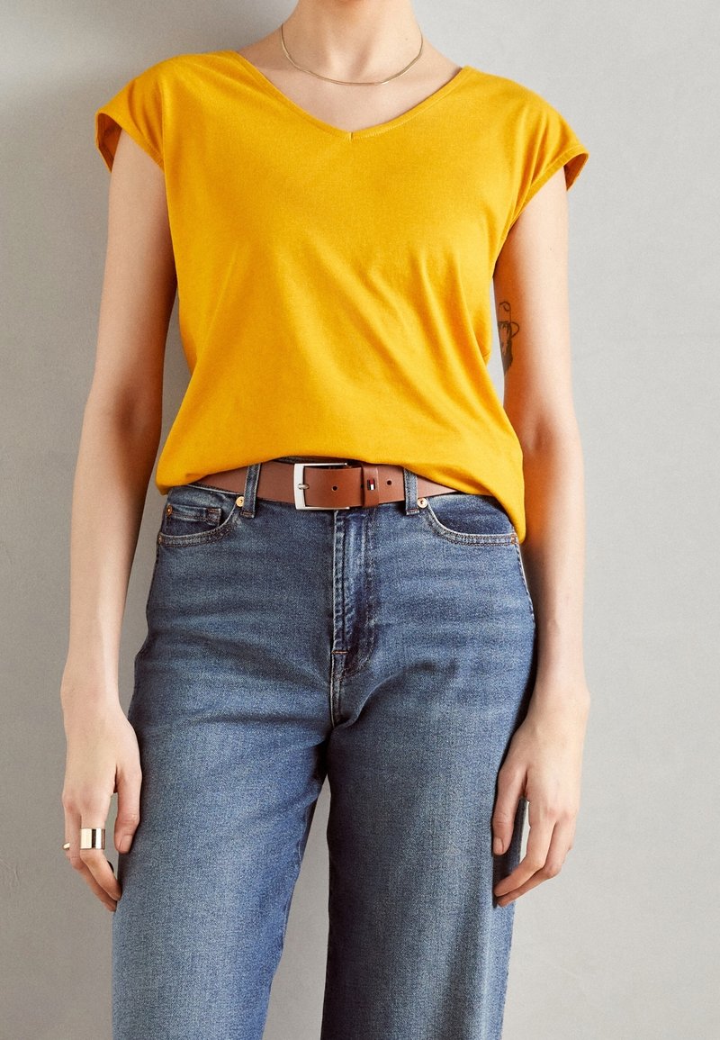 Person som bär en senapsgul kortärmad topp stoppad i högmidjade blå jeans med ett brunt bälte och minimal smyckesdesign.