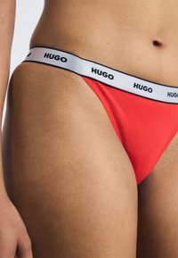 Red thong underwear with a white waistband featuring the brand name "HUGO" in black. Smooth fabric with a form-fitting design.