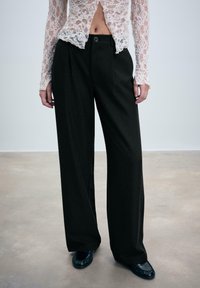 Eine Person trägt schwarze weite Hosen, schwarze Schuhe und ein weißes langärmliges Spitzenshirt. Sie steht auf einem glatten Boden vor einem schlichten Hintergrund.