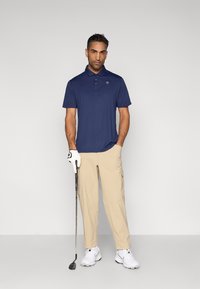 Μπλε ναυτικό πόλο με κοντά μανίκια και ραφτή λωρίδα με κουμπιά, συνδυασμένο με ανοιχτό μπεζ παντελόνι. Τα αξεσουάρ περιλαμβάνουν ένα λευκό γάντι γκολφ και ρόπαλο.