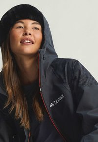 Svart och grå vattentät jacka med huva fodrad med päls, utrustad med kontrasterande texturerade axelpaneler och en framträdande logotyp på bröstet.