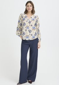 Hellbeige Bluse mit blauem Blumenmuster, weichem V-Ausschnitt und Puffärmeln, kombiniert mit marineblauen Hosen mit weitem Bein und spitzen Schuhen.